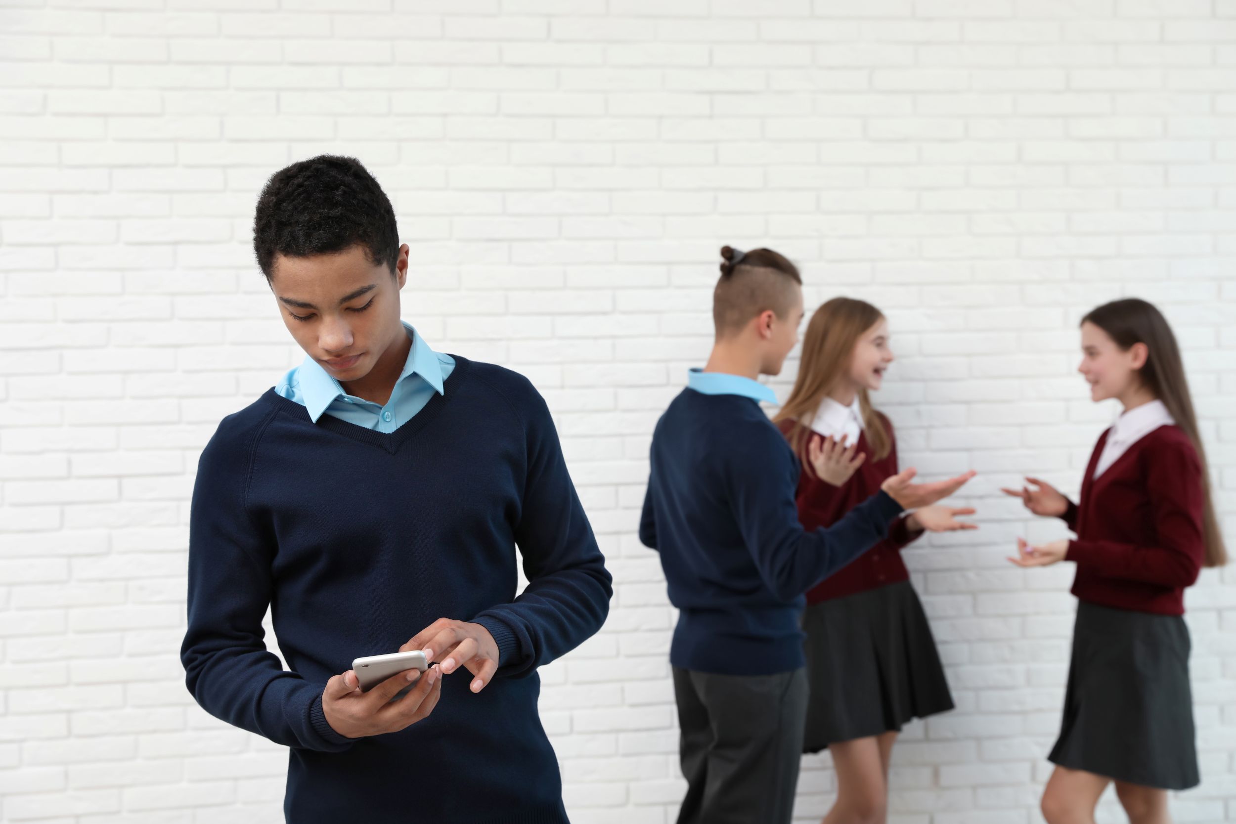 A teenage student stands alone looking down at his mobile. Behind him, three other students stand chatting and laughing