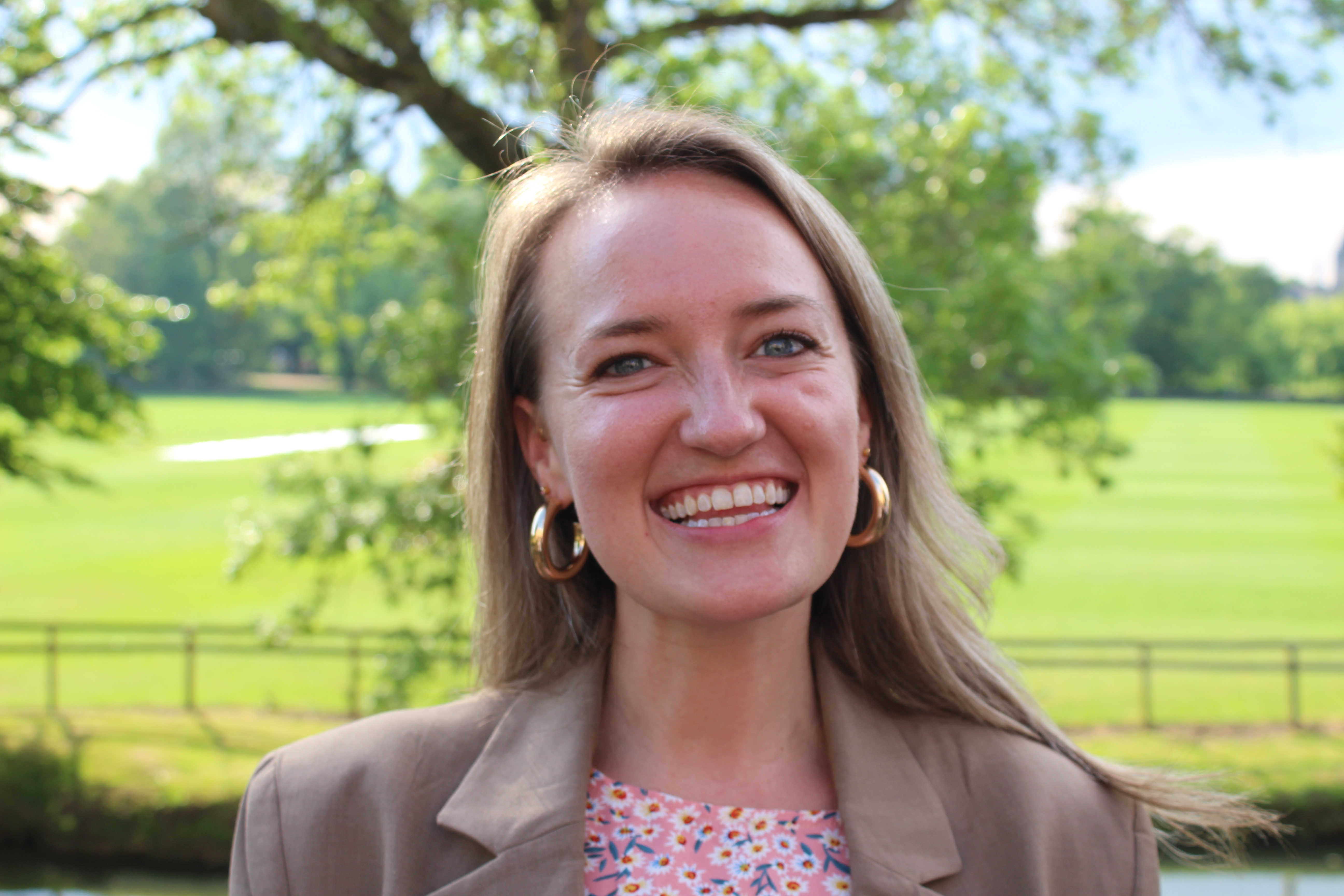 A smiling woman outdoors in a sunlit green park, wearing gold hoop earrings, a taupe blazer, and a pink-and-white patterned top. Trees and an open grassy field are softly blurred in the background.