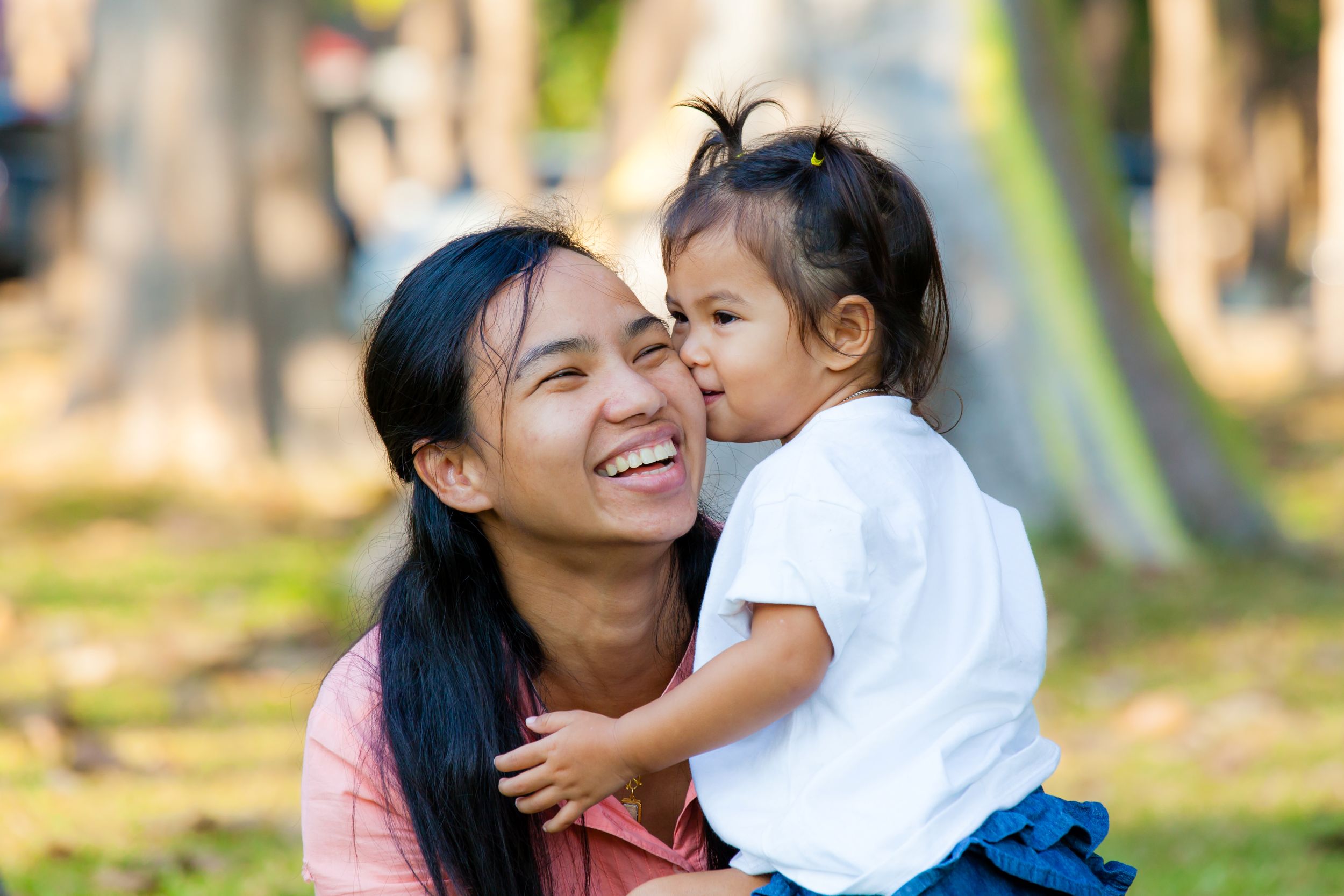 A mother crouching down next to her young daughter, who is kissing her check. The mother is laughing. 