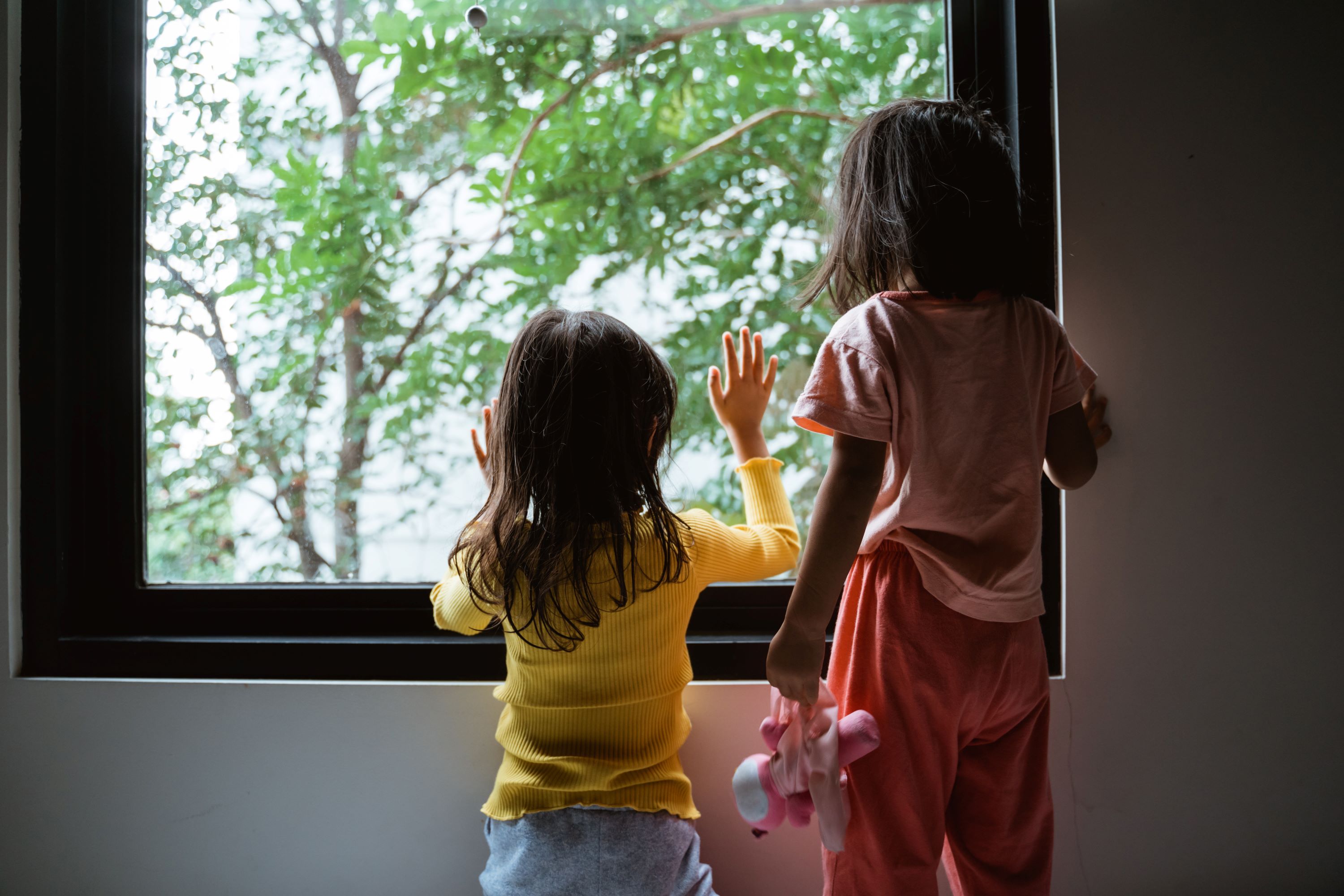 Two young children seen from behind. They are standing and looking out of a large window at some trees. -- Odua Images, stock.adobe.com