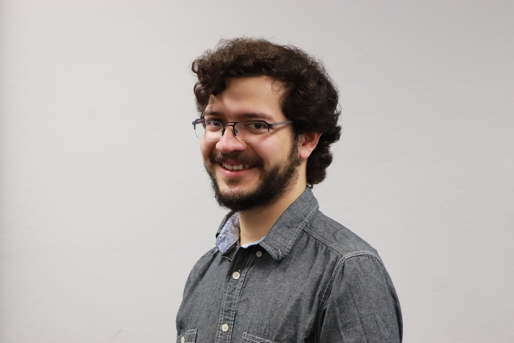Man with curly dark hair and beard and wearing dark rimmed glasses and grey shirt