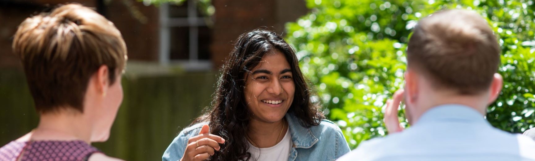 Three people sat outside in the sunshine around a table. Two of the people have their back to the camera on the near side of the table. The student facing the camera is on the other side, wearing a denim shirt and white tee. She is smiling. 