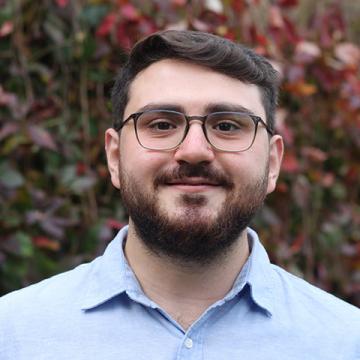Ertugrul has short brown hair, a brown beard and glasses. He's wearing a pale blue shirt