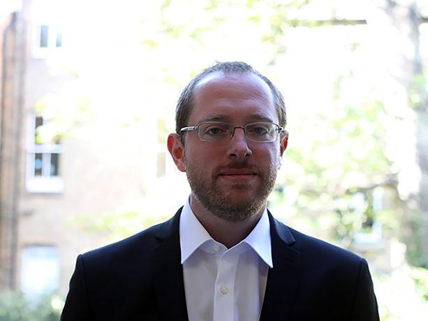 Man with light brown hair and gold rimmed glasses, wearing a white shirt and dark suit