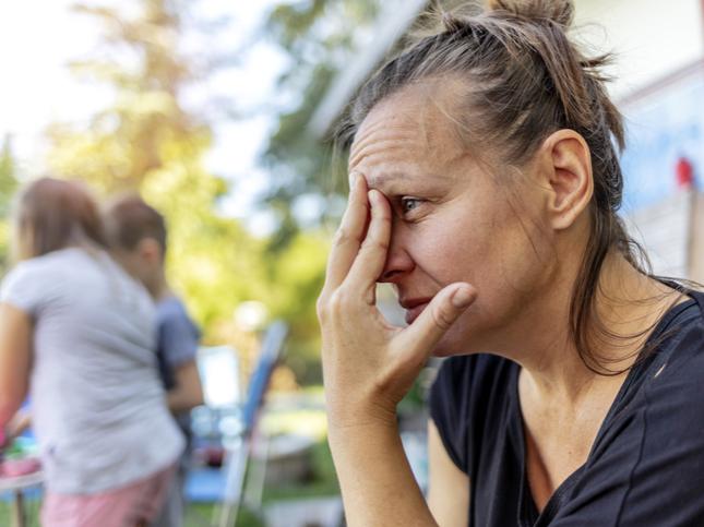A mother sitting outside, resting her forehead on her fingertips. Children are playing behind her. 