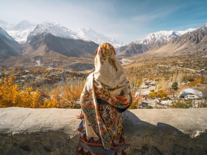 A woman wearing traditional dress sitting on wall and looking at Hunza valley in autumn season, Gilgit Baltistan in Pakistan, Asia