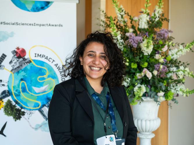 Mona Ibrahim has long curly dark hair. She is smiling broadly, standing in front of a bouquet of flowers and an 'Impact Awards' banner