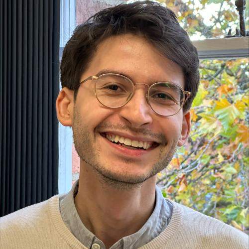 David has short dark hair, is wearing glasses and a cream coloured jumper. There is a window behind him with autumnal trees outside.
