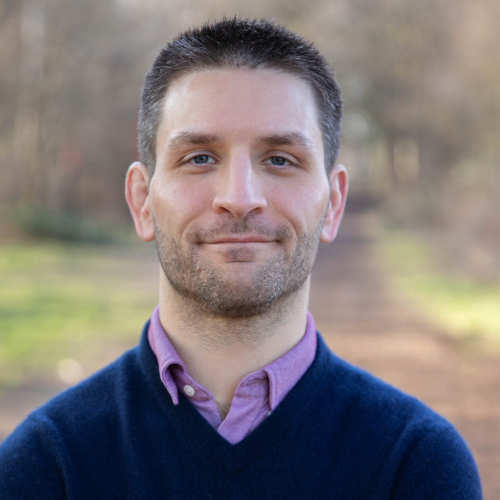 Lewis Anderson is wearing a dark blue sweater over a purple shirt, with short dark hair, looking directly at the camera.