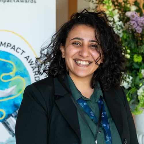 Dr Mona Ibrahim has shoulder-length curly black hair. She is smiling broadly, wearing a dark green blouse and black blazer.