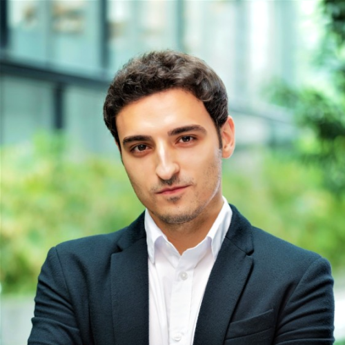 Alessio Cuozzo has short dark hair. He is wearing a white shirt and navy blazer, and is standing in front of a glass building.
