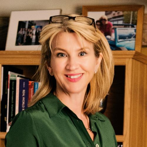 Caroline Greenhalgh wears a dark green shirt, has shoulder-length blonde hair and stands in front of a bookshelf. 