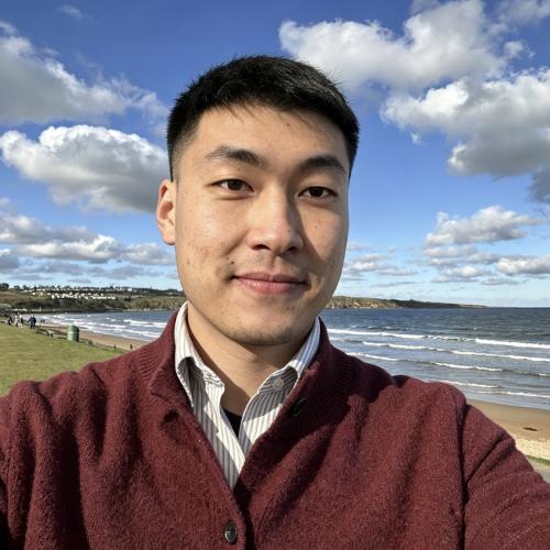 Man with short dark hair, wearing a jumper and standing near the sea