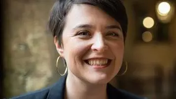 Dr Aoife O’Higgins is wearing a navy blue jacket and top. She has short dark hair, large hoop earrings and is smiling at the camera 