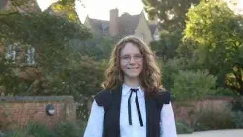 Nicola Tomlin wearing full academic dress and is standing in a college garden.  Nicola has long brown curly hair and wearing glasses