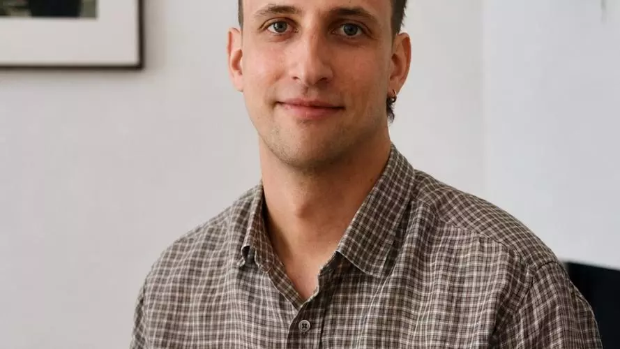 Man with short brown hair wearing a brown checkered button-up shirt, seated indoors against a white wall with framed artwork and a leafy plant in the background