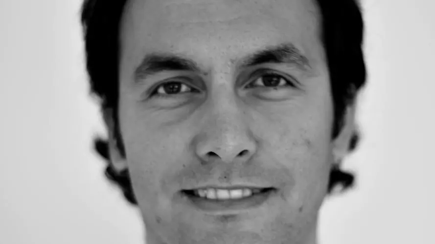 A black and white photo of Erdem Yörük, who has curly dark hair and is wearing an open-collared white shirt