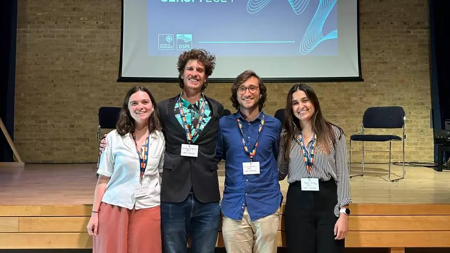 A group of four students standing with their arms around each other, in front of a raised stage and display screen that says 'SERSPI 2024'.