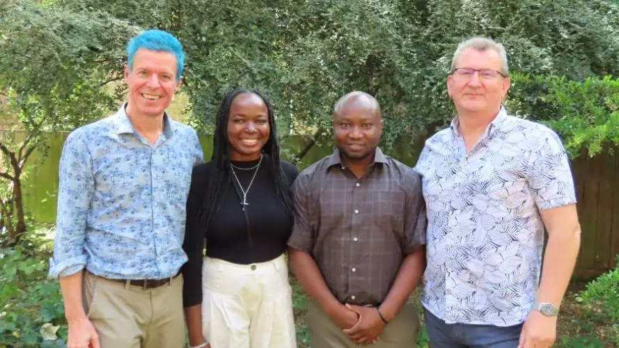 Four people smiling outdoors in front of greenery, standing side by side smiling at the camera.