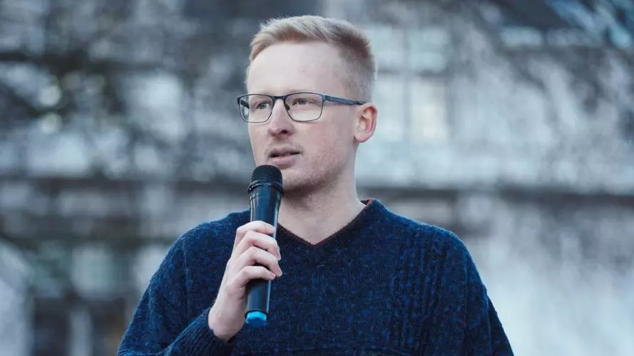 In jeans and a dark blue top, Ben Goodairds is holding a microphone and appears to be speaking outside in a public space.
