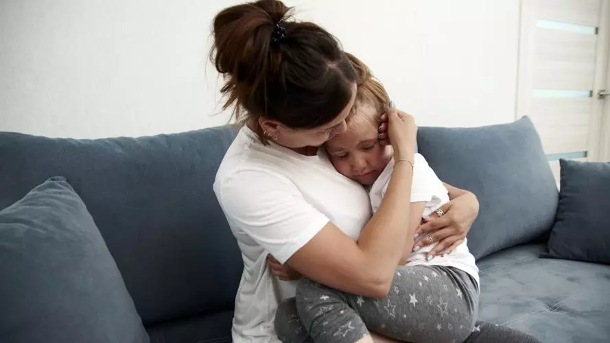 A mother sits on a grey sofa, hugging and comforting her young daughter