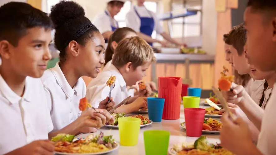 Several primary school children sitting at a lunch table, wearing white polo shirts. They are eating and chatting. The table is filled with multicoloured cups.