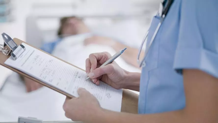 A close up of a nurse writing notes on a clipboard. In the background is a patient in a hospital bed.