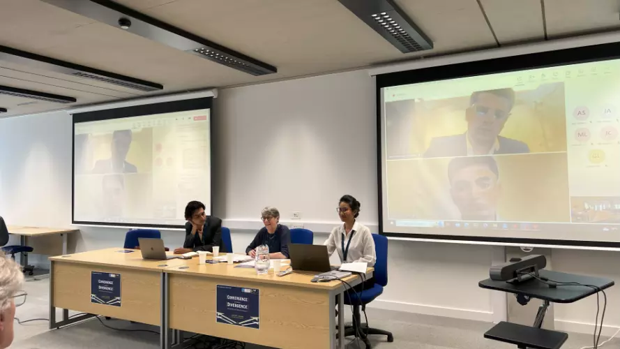 A panel session is taking place in a room, with 3 people sitting in a room with two white screen appearing to be giving a presentation.