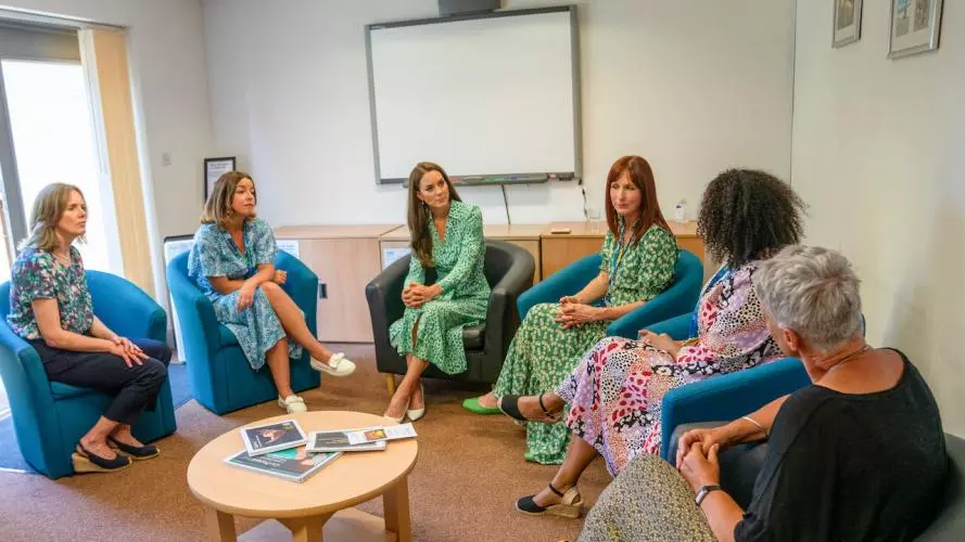  Several women, having a discussion, are sitting on chairs arranged in a semi-circle, with a projector screen in the background