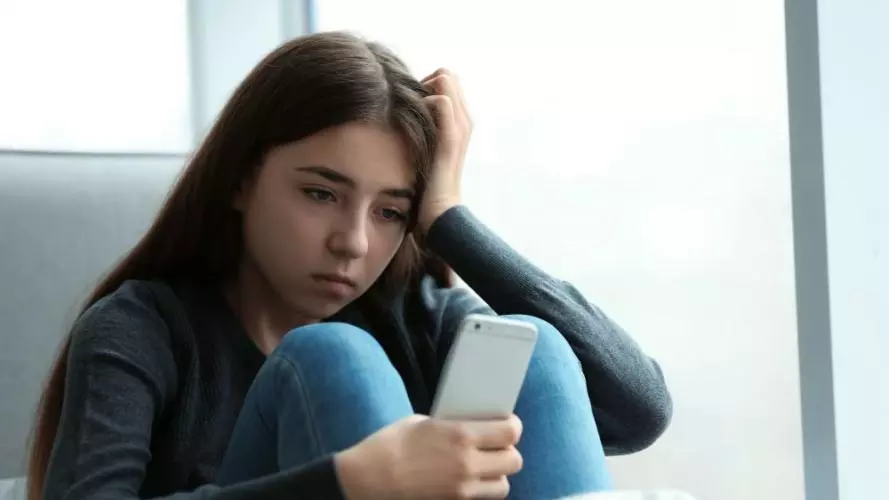 A young girl with long dark hair is sitting on a chair, looking at her phone with a sad expression. 