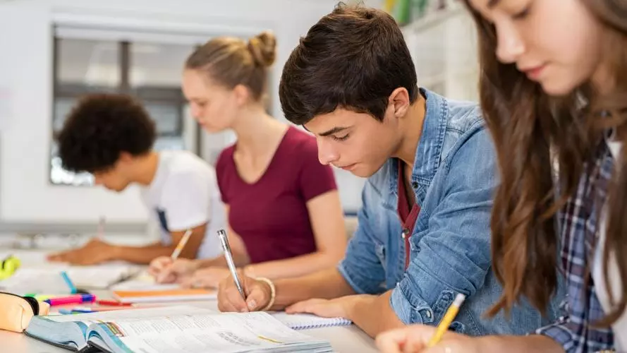 Four young adult students sat in a row at a desk, holding pens and looking at textbooks. 