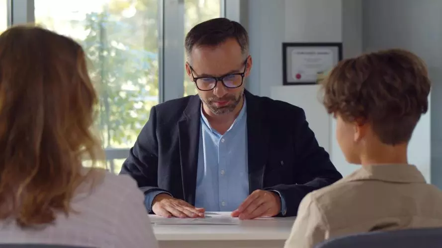A male teacher sitting at a desk, talking to a mother and child who are sat opposite him with their backs to the camera.