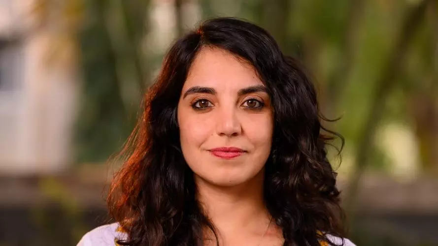 Wearing a pink top with mustard flower patterns and shoulder-length brown wavy hair, Anita smiles gently at the camera.
