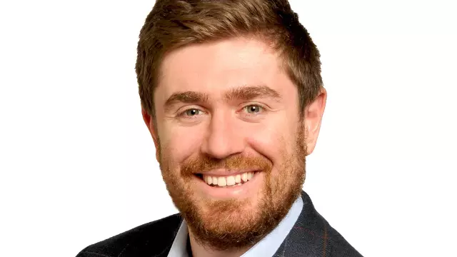 Benjamin Scher has short ginger hair and a beard, wearing a blue shirt with a dark blue suit jacket, and smiling at the camera.