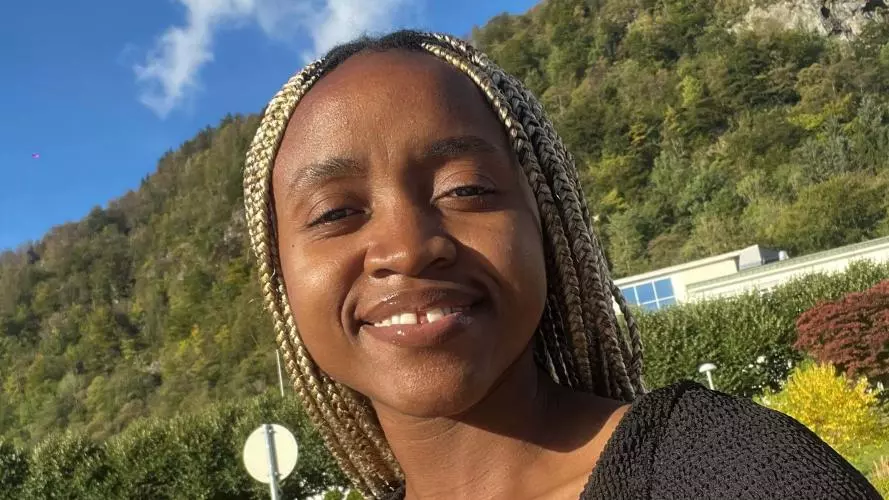 Diana Mwala is smiling in front of a blue sky. Her hair is braided and there is a tree-lined hill in the background. 