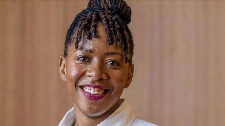 Dr Nontokozo Langwenya wears a white shirt and is smiling in front of a wood-panelled background.