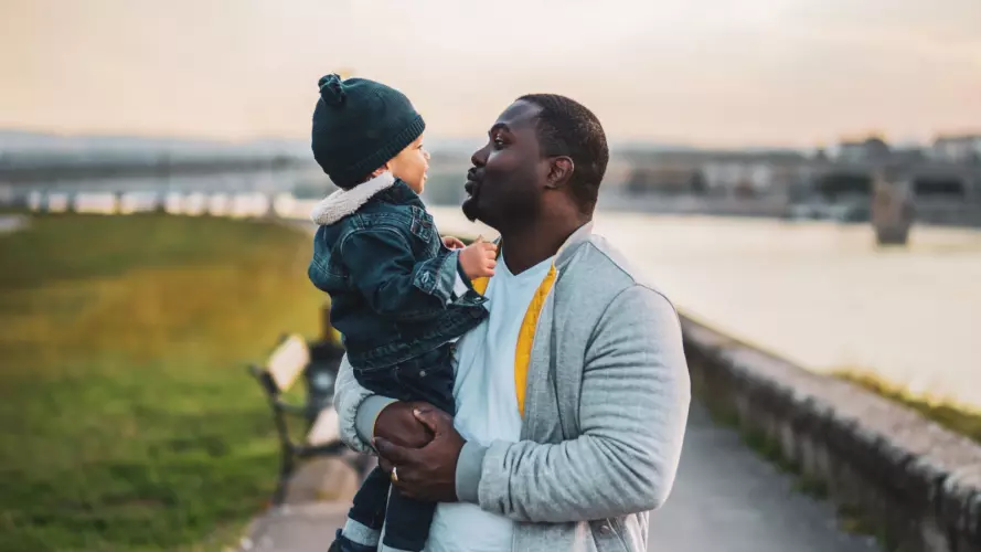 A father A holding a baby boy, with a river in the background.