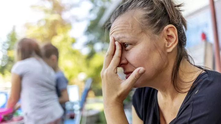 A mother sitting outside, resting her forehead on her fingertips. Children are playing behind her. 