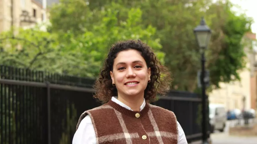 Elifcan has shoulder-length, curly dark hair. She is wearing a white shirt and a brown vest with a large white check. She is standing in front of a park fence, smiling. 