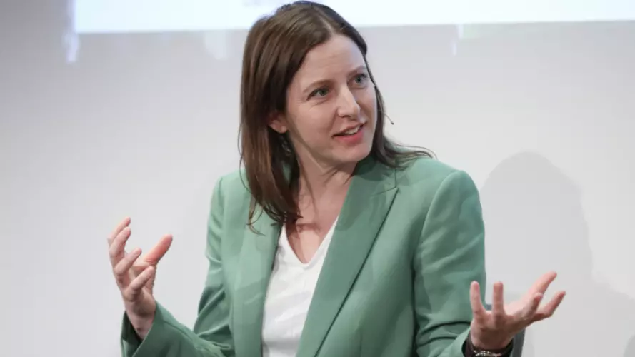 As Silja Häusermann sits on a stage wearing a green suit, she appears to be in a discussion and expressing herself with her hands. 
