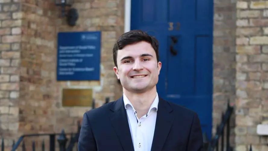 Isaac Mellis-Glynn Stands in front of the DSPI building. He has dark brown hair and wears a white shirt and navy blazer.