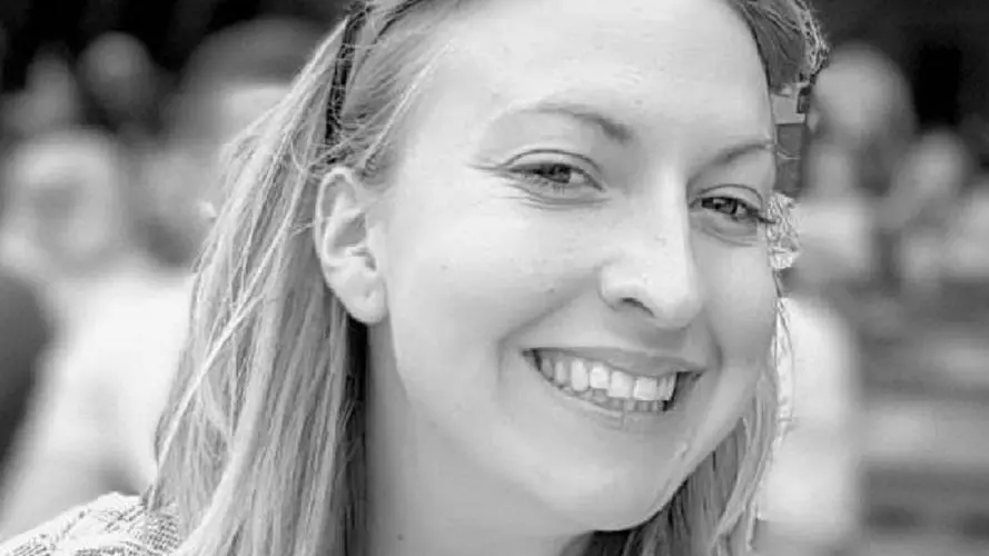 kathryn has shoulder-length blonde hair that is held back from her face by glasses on her head. She's smiling at the camera and is wearing a checked, woven jacket 