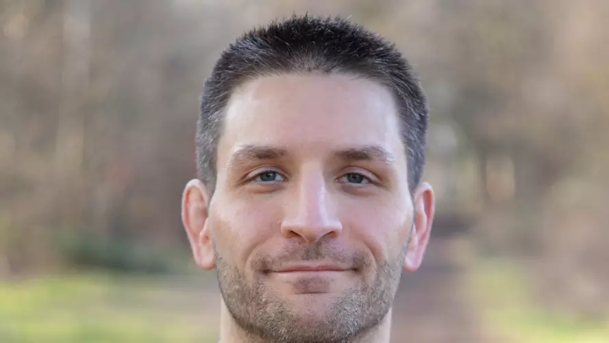 Lewis Anderson is wearing a dark blue sweater over a purple shirt, with short dark hair, looking directly at the camera.
