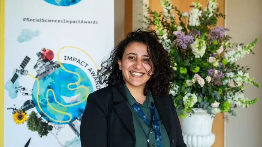 Mona Ibrahim has long curly dark hair. She is smiling broadly, standing in front of a bouquet of flowers and an 'Impact Awards' banner