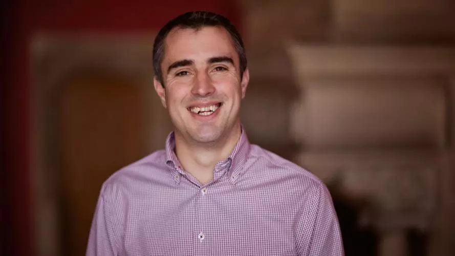 Peter Burr is wearing a purple shirt, with short dark hair and is smiling at the camera.