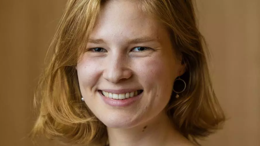 Sara Hueber has bobbed ginger hair and is wearing a white top. She is in front of a brown background, smiling. Credit: Tom Weller Photography, 2025