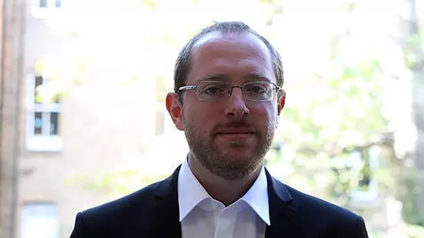 Man with light brown hair and gold rimmed glasses, wearing a white shirt and dark suit