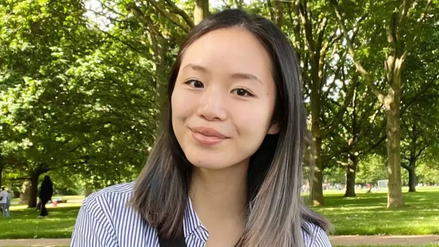 Victoria Tan has shoulder length dark hair, wearing a blue and white top and is sitting in a park.
