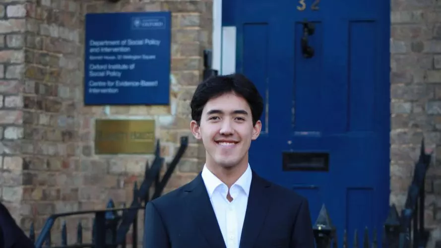 Xin Law-Gallagher stands, smiling, in front of the DSPI blue door. He wears a white shirt and dark blazer. He has short cropped dark hair. 