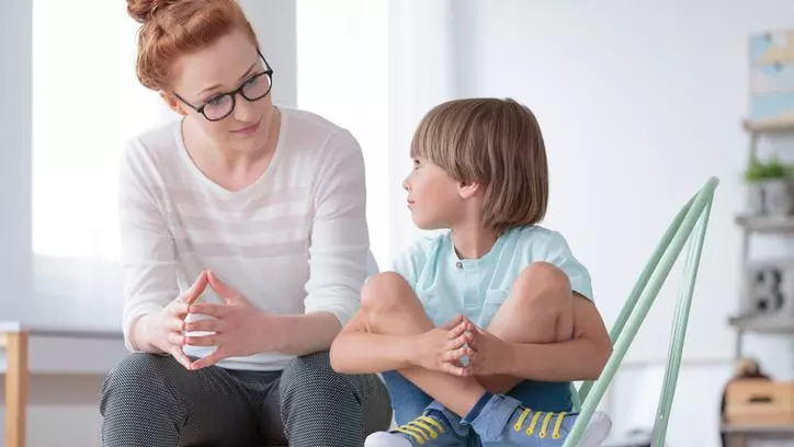 A young boy sits cross-legged on a chair, looking up at an adult woman sat next him.